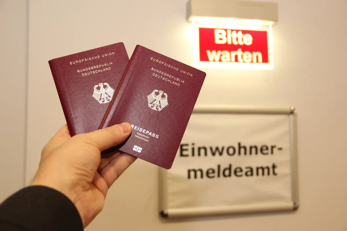 Eine Hand hält zwei Reisepässe. Im Hintergrund ist ein Schild mit der Aufschrift "Einwohnermeldeamt" zu sehen.