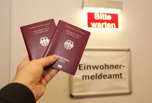 Eine Hand hält zwei Reisepässe. Im Hintergrund ist ein Schild mit der Aufschrift "Einwohnermeldeamt" zu sehen.
