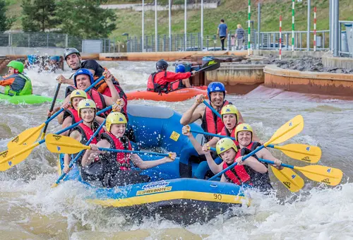 Ein Schlauchboot mit neun Rafting-Teilnehmern fährt den Wildwasserkanal hinab. Im Hintergrund tarinieren Kajaksportler.