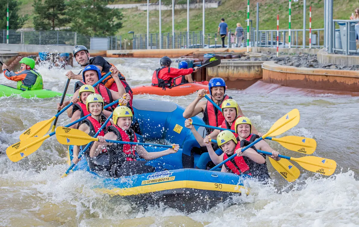 Ein Schlauchboot mit neun Rafting-Teilnehmern fährt den Wildwasserkanal hinab. Im Hintergrund tarinieren Kajaksportler.