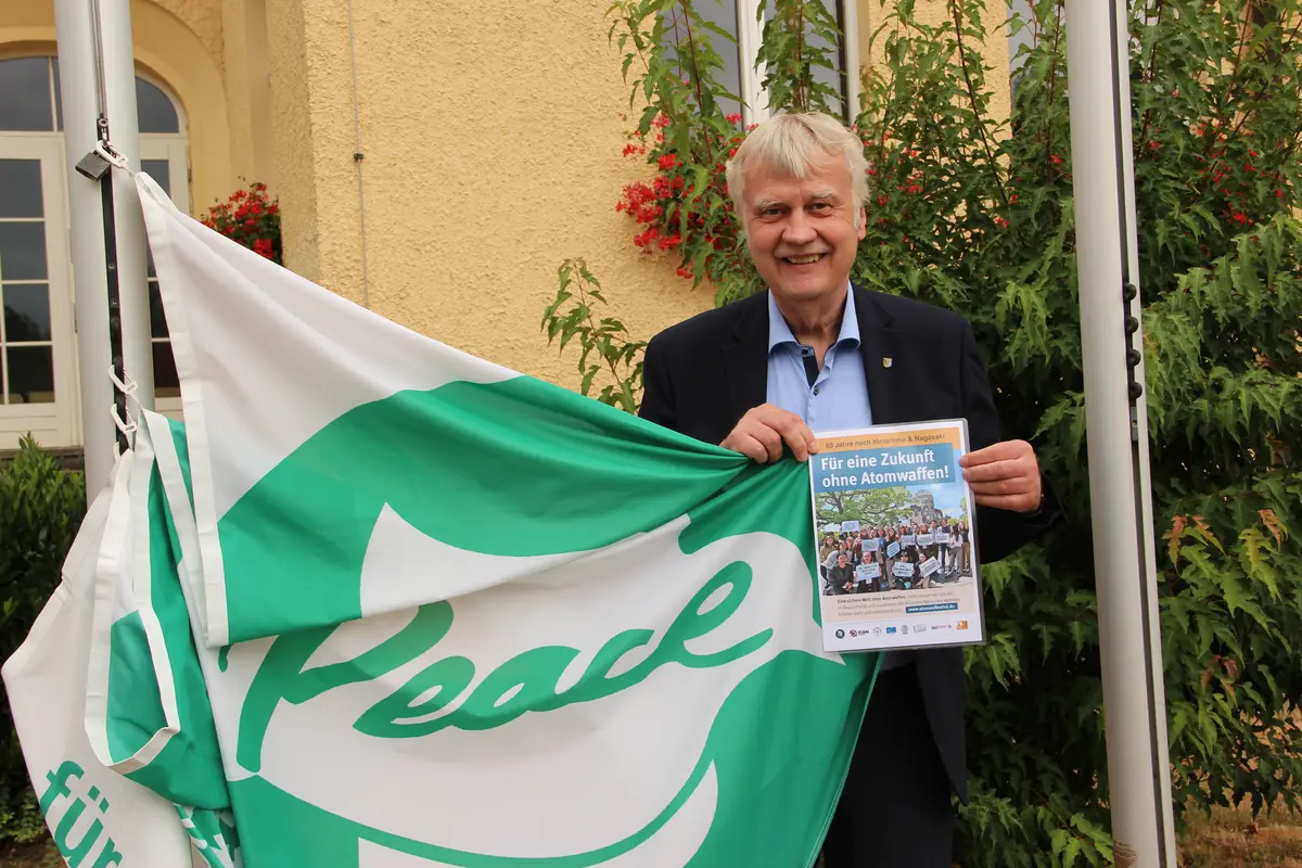 Oberbürgermeister Karsten Schütze hält die Flagge der "Mayors for Peace" so, dass der Schriftzug "Peace" zu sehen ist.